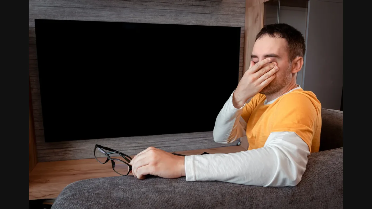Frustrated man covering his face in front of a large black, blank television screen