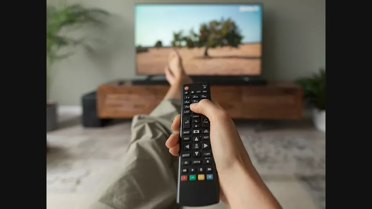 Hand holding a TV remote in front of a television, with feet up on a sofa