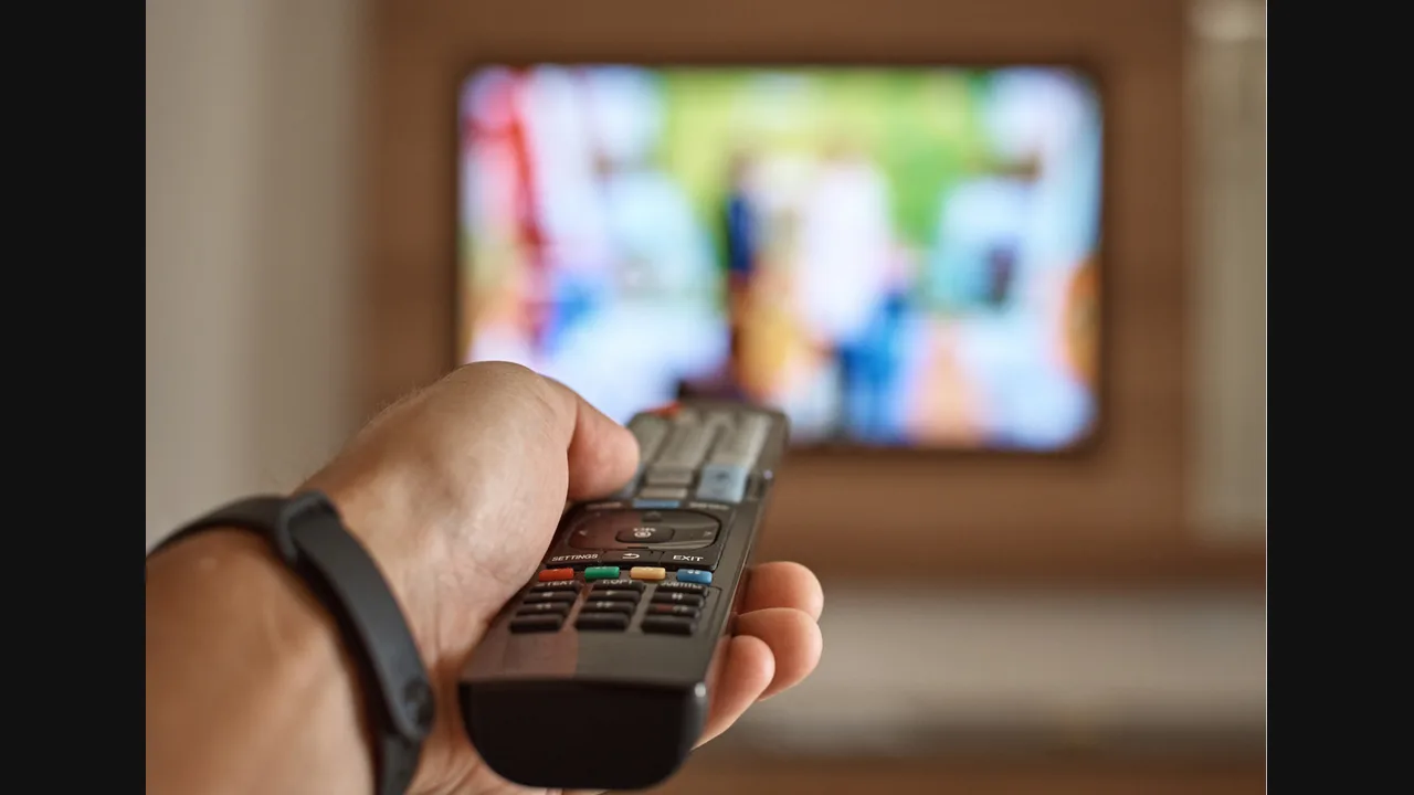 Hand holding a TV remote pointed at a blurred television screen